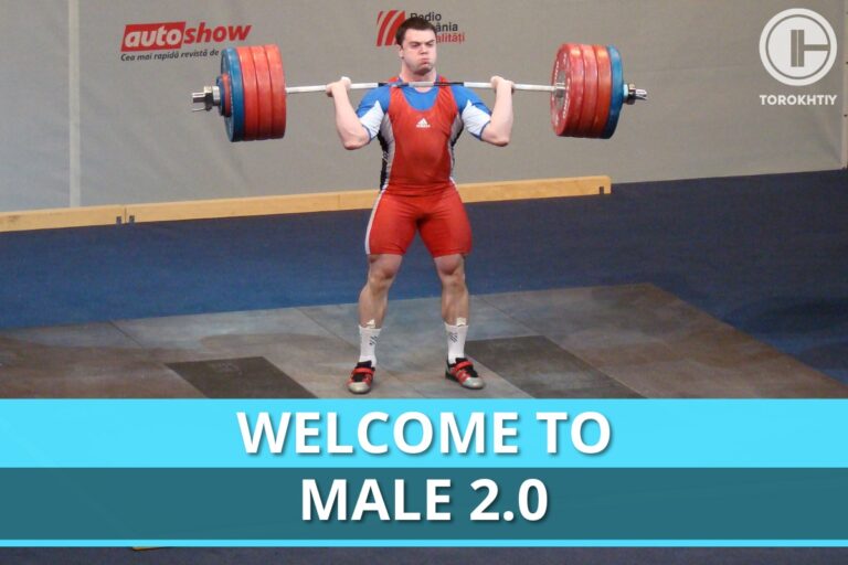 A male weightlifter in a red uniform holds a barbell at shoulder height during competition, with Welcome To Male 2.0 banner behind him. (Torokhtiy Weightlifting)