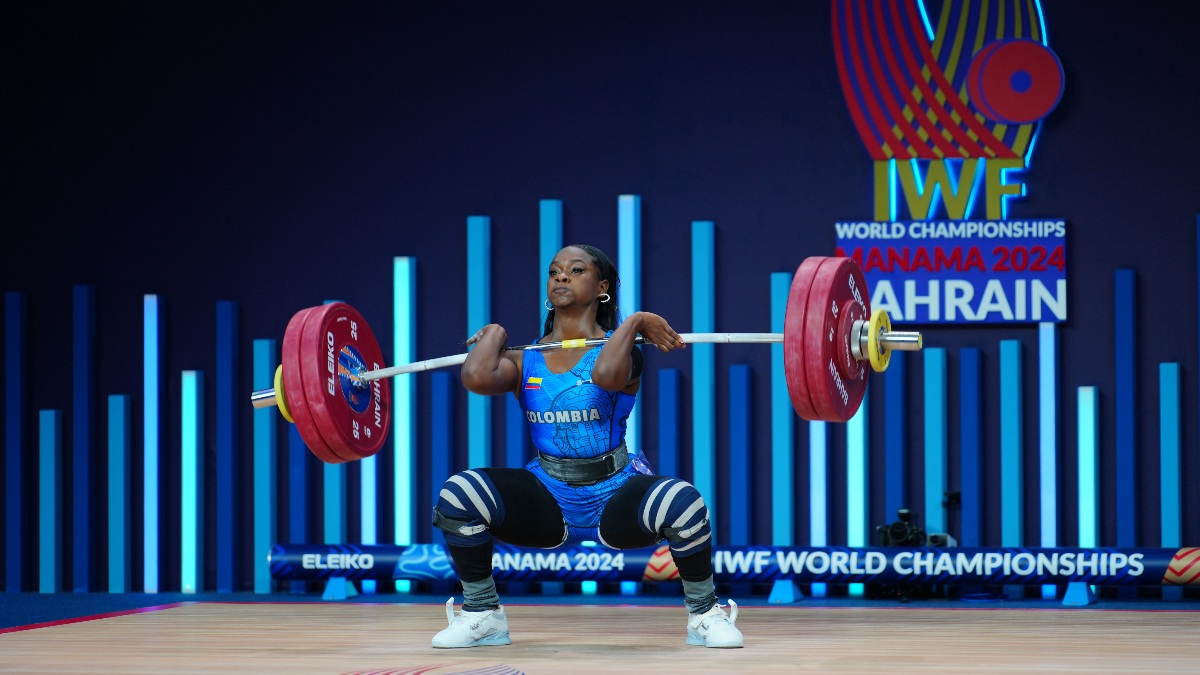 Colombian weightlifter Vanesa Segura lifts a barbell at IWF World Championships Bahrain 2024, event logo and blue backdrop behind. (Torokhtiy Weightlifting)