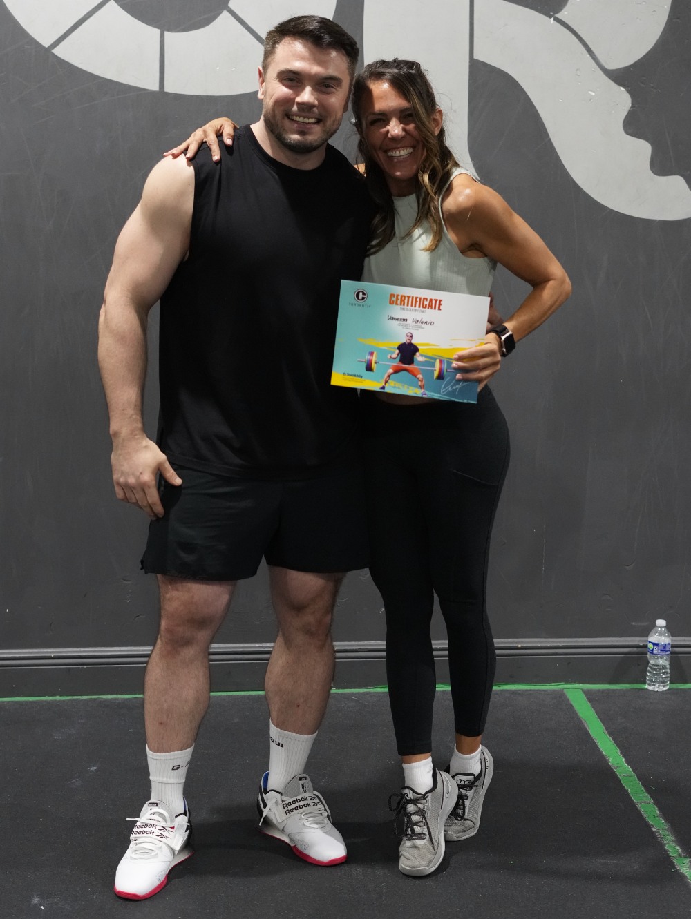 After a fitness seminar, two athletes pose in gym wear as one proudly holds a certificate; dark wall and water bottle behind them. (Torokhtiy Weightlifting)